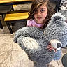 child, teddy_bear, toy, indoor, kitchen, table, bench, tile_floor, curly_hair, pink_shirt, stuffed_animal, person, looking_up, plush, home, furniture, carpet, cute, young, holding