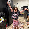 Callie joined the competition — help win amazing prizes! child, barefoot, kitchen, refrigerator, stove, decorative_item, rug, tile_floor, tshirt, shorts, pet_bowls, trash_can, table, chair, calendar, floor_mat, playful, person, indoor, home