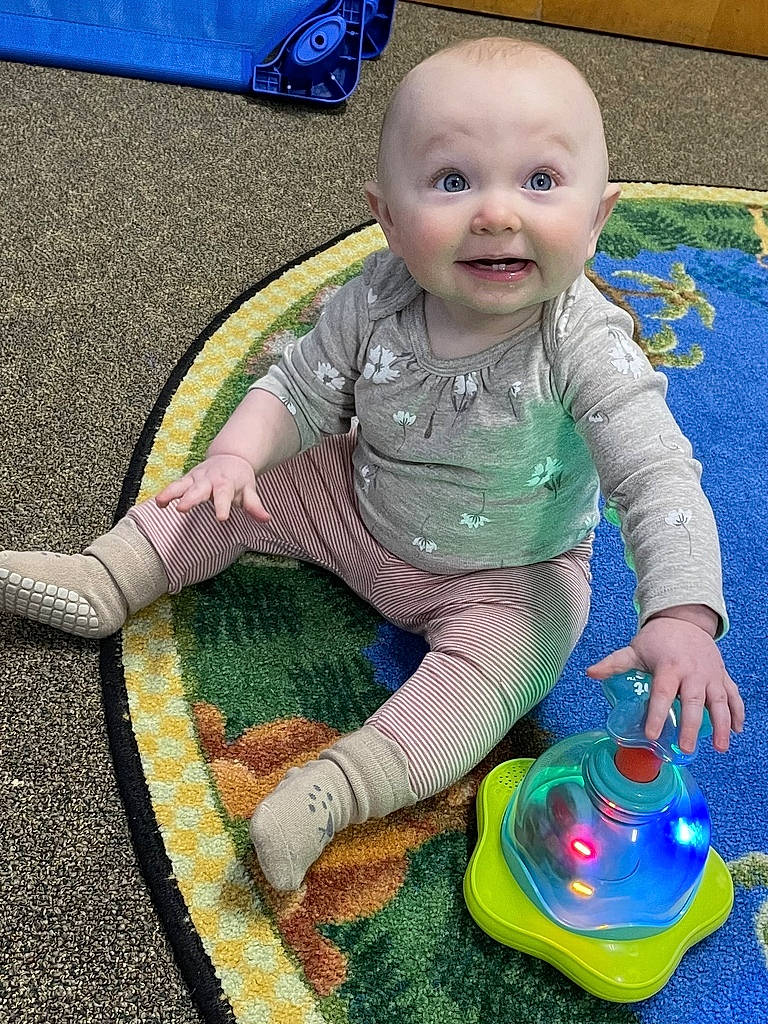 Lucy is registered to the contest to win money with this photo: baby, baby_products, baby_toddler_clothing, baby_toys, ball, blue, cheek, child, circle, electric_blue, face, flooring, fun, grass, happy, joy, pattern, person, play, sitting