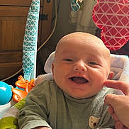 George is registered to the contest to win money with this photo: baby, infant, smiling, happy, play_seat, toys, hand, clothing, face, indoor, furniture, colorful, cute, child, person, portrait, cheek, finger, green, fabric