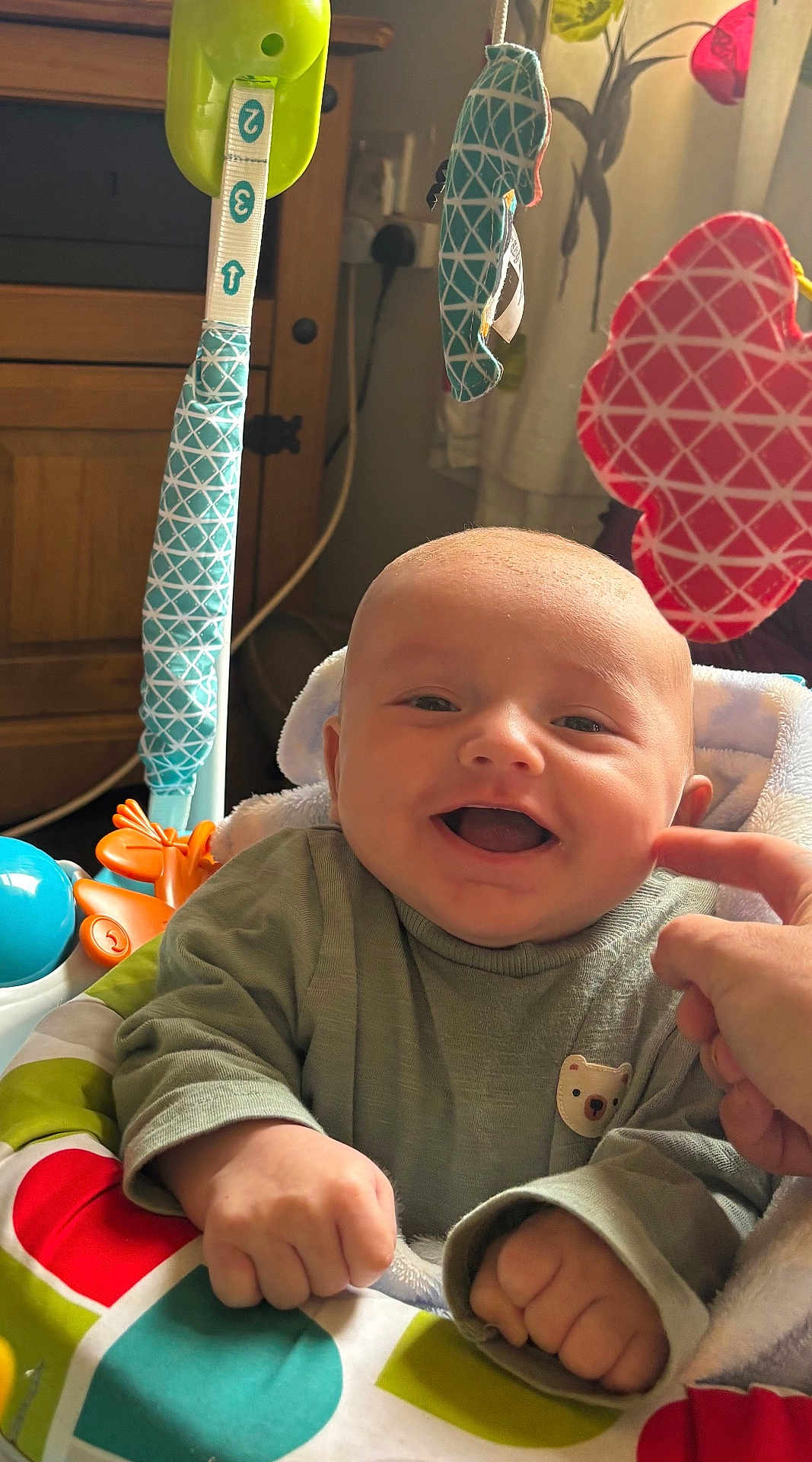 George is registered to the contest to win money with this photo: baby, infant, smiling, happy, play_seat, toys, hand, clothing, face, indoor, furniture, colorful, cute, child, person, portrait, cheek, finger, green, fabric