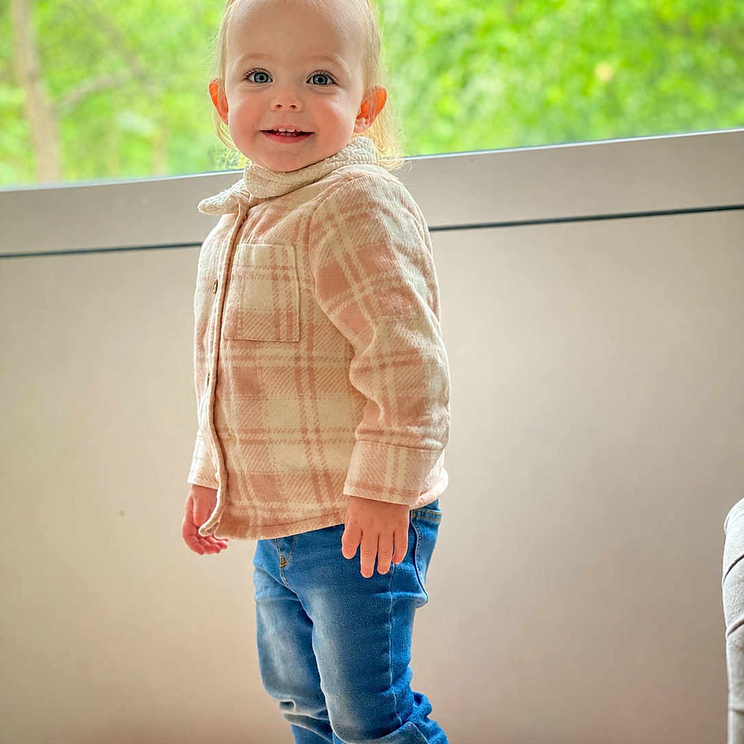 Skylar-Rose is registered to the contest to win money with this photo: toddler, child, standing, indoors, jeans, jacket, smiling, happy, window, greenery, face, footwear, casual_clothing, blond_hair, portrait, young_child, natural_light, cozy, cute, person