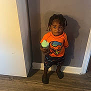 Chozen joined the competition — help win amazing prizes! child, toddler, person, standing, orange_shirt, shorts, holding, cup, indoors, floor, wood_floor, wall, shadow, hair, braids, feet, footwear, casual_clothing, expression, room