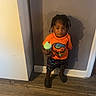 child, toddler, person, standing, orange_shirt, shorts, holding, cup, indoors, floor, wood_floor, wall, shadow, hair, braids, feet, footwear, casual_clothing, expression, room