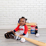 Kyrie is registered to the contest to win money with this photo: baby, baseball, bat, glove, ball, floor, crate, snacks, coca_cola, brick_wall, wooden_floor, child, smiling, sitting, sports_equipment, playful, portrait, indoor, clothing, person