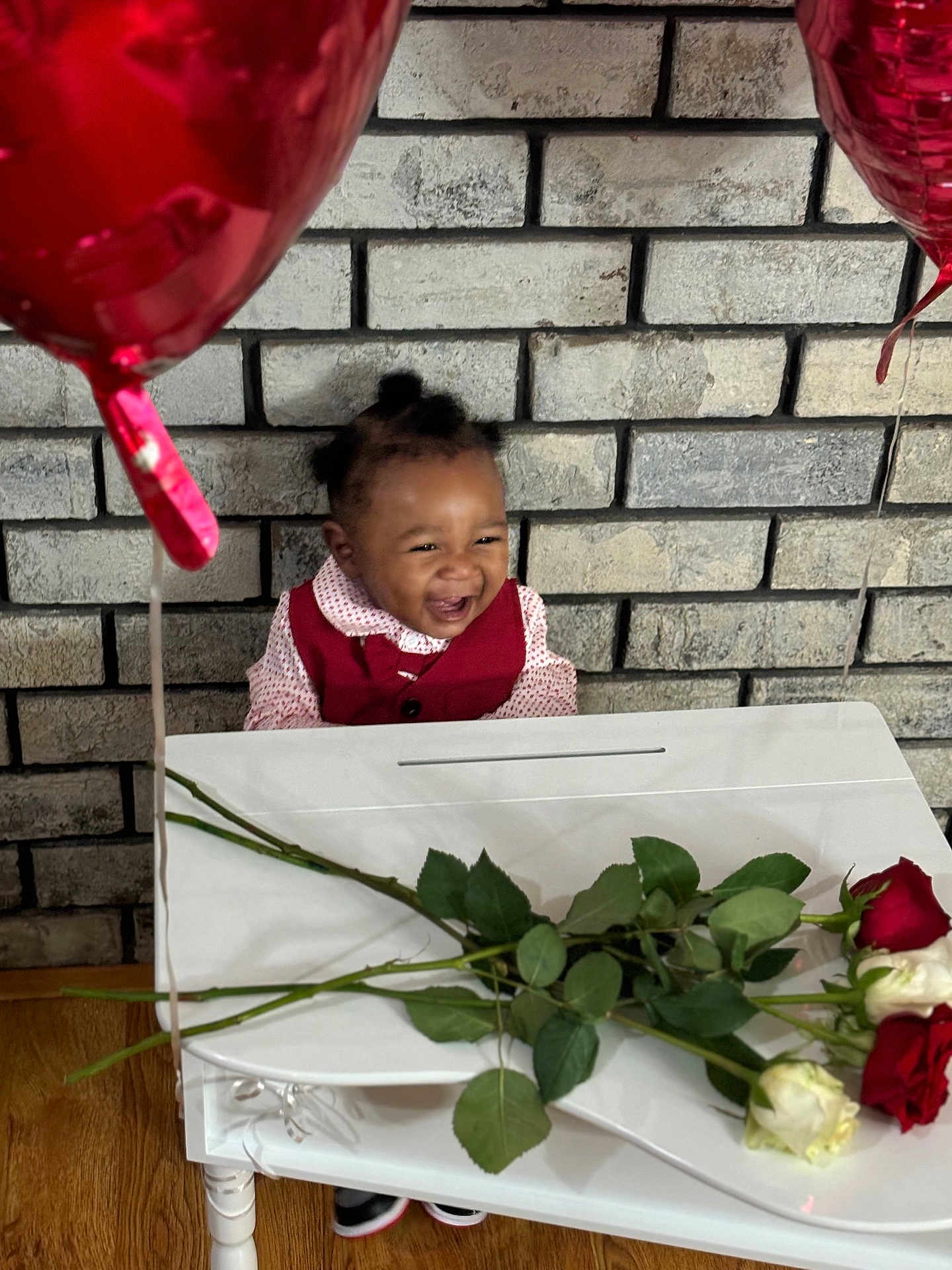 Kyrie joined the competition — help win amazing prizes! toddler, smiling, child, red_vest, white_shirt, balloons, roses, table, brick_wall, wooden_floor, happy, celebration, indoors, plant, flower, person, portrait, cute, decor, joy