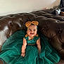 baby, child, smiling, dress, headband, bow, green_dress, gold_accents, couch, leather, indoor, happy, sitting, cute, portrait, plush, tulle, fashion, person, infant