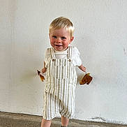 Liam participe au concours pour gagner de l'argent avec cette photo : toddler, child, blonde_hair, blue_eyes, striped_overalls, white_shirt, sandals, leaves, autumn, standing, smiling, concrete_floor, plain_wall, casual_clothing, happy, playful, portrait, cute, indoor, young_child