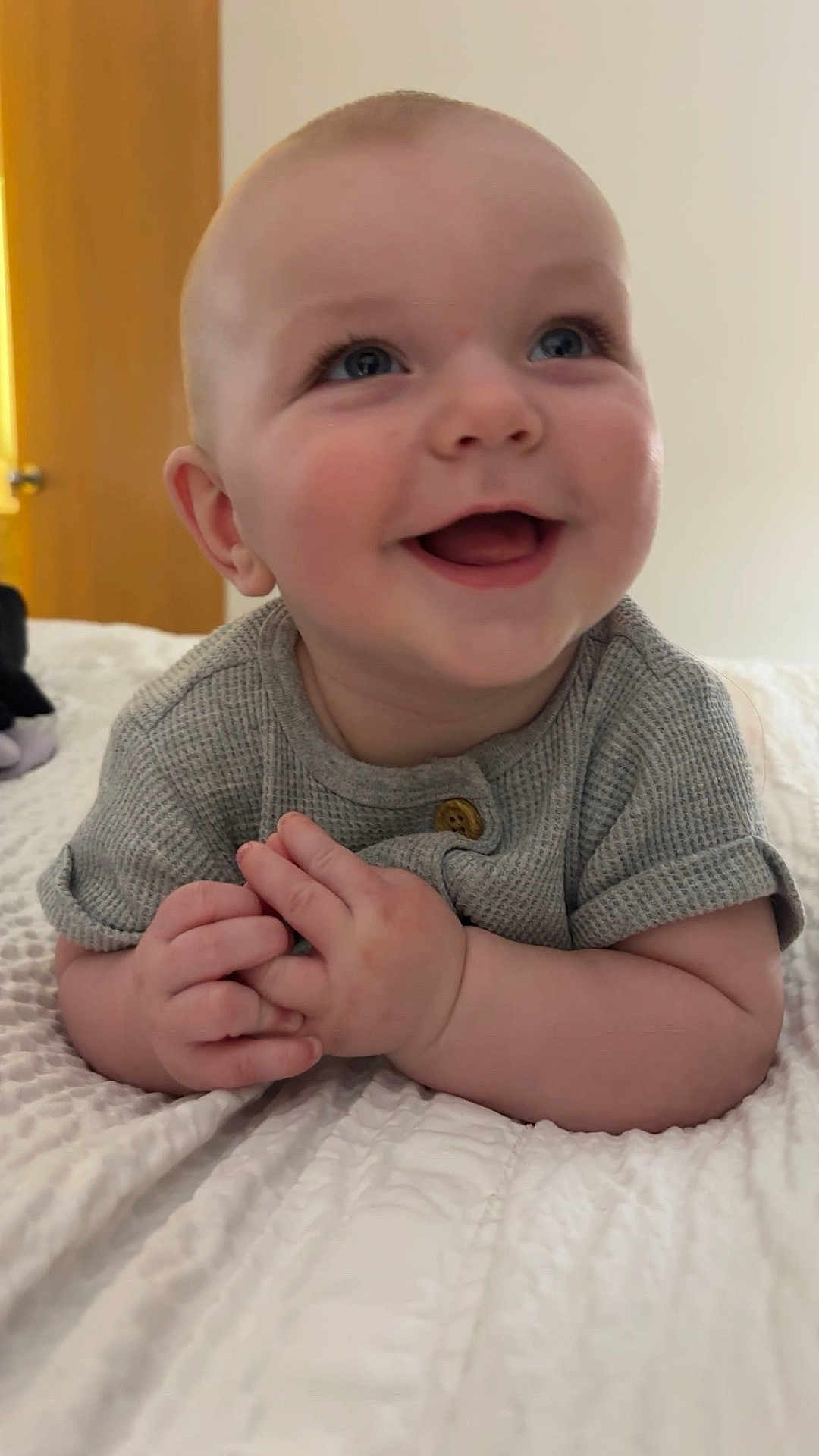 John joined the competition — help win amazing prizes! baby, infant, smiling, happy, face, hands, blue_eyes, short_hair, gray_shirt, bed, blanket, indoor, cute, portrait, child, skin, young, person, expression, lying_down