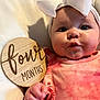 Ariella is registered to the contest to win money with this photo: baby, infant, child, face, headband, bow, clothing, pink, hand, wooden_sign, text, blanket, soft, portrait, closeup, cute, skin, eyes, expression, indoor