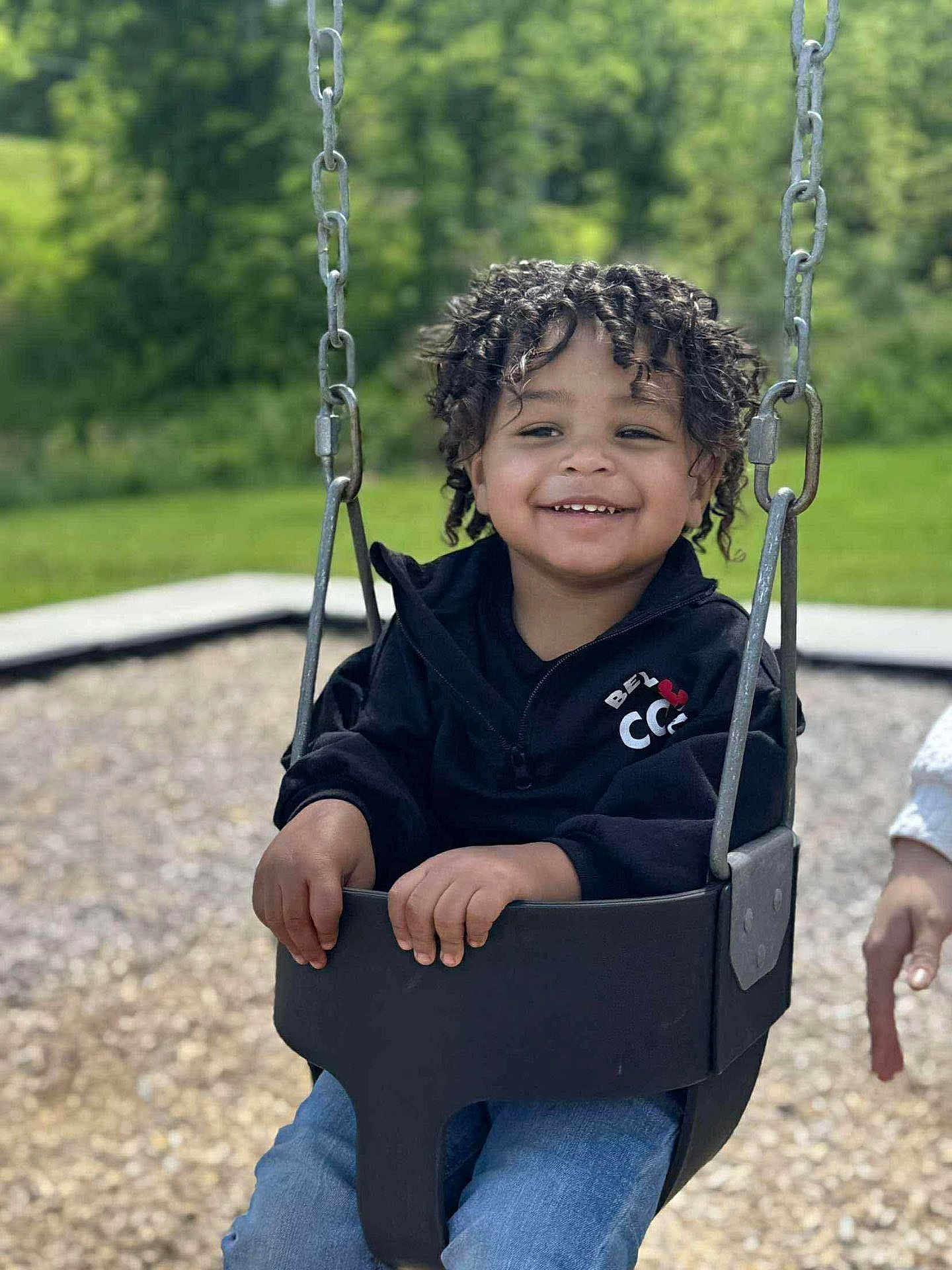 Dior is registered to the contest to win money with this photo: black_jacket, casual_clothing, child, curly_hair, daylight, fun, greenery, happy, holding, jeans, metal_chains, outdoor, park, person, play, playground, seat, smiling, swing, toddler