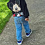 back_view, casual_clothing, child, curly_hair, cute, daylight, fashion, grass, jeans, kid, mickey_mouse, outdoor, pavement, person, sidewalk, sneakers, sweatshirt, urban, walking, young