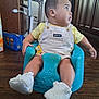 Onyx is registered to the contest to win money with this photo: baby, child, infant, blue_seat, yellow_shirt, white_overalls, pacifier, baby_booties, wooden_floor, kitchen, cabinet, box, curious, sitting, indoor, person, looking_away, home, furniture, floor