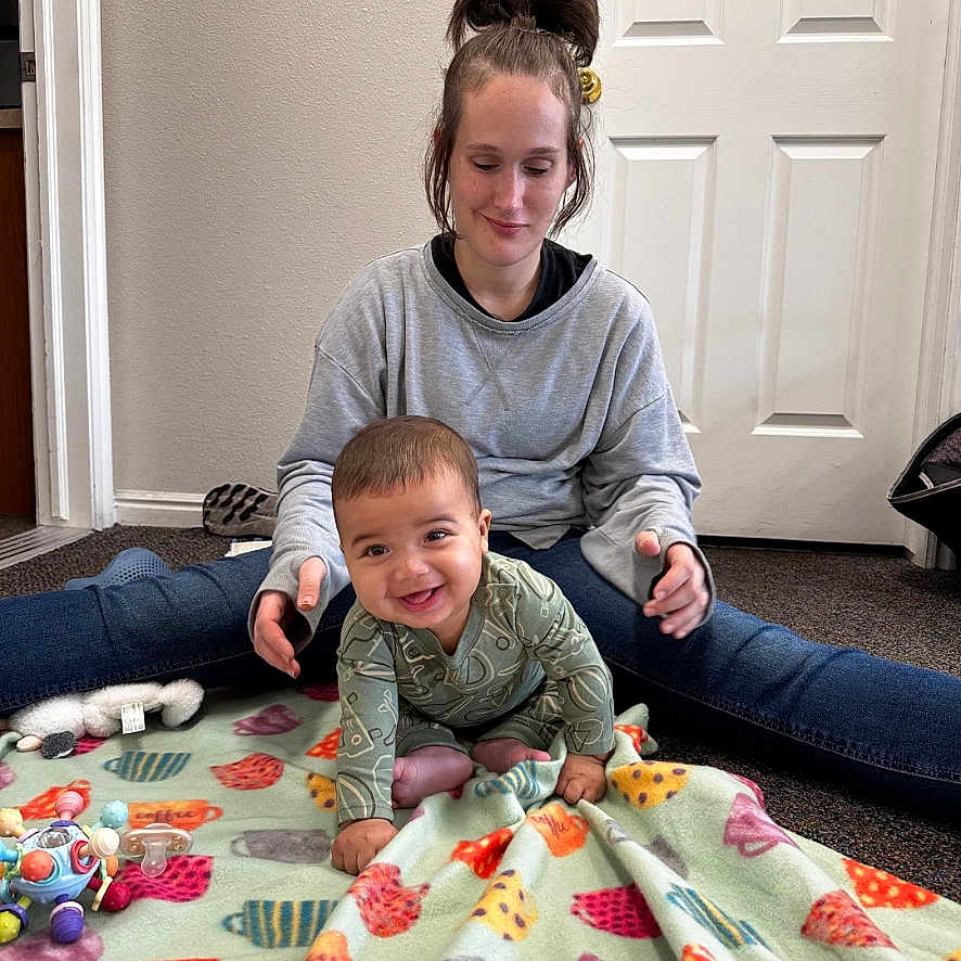 Onyx is registered to the contest to win money with this photo: baby, blanket, carpet, casual_clothing, child, crawling, cute, door, family, happy, home, indoor, person, playing, relaxed, sitting, smiling_baby, toys, woman, young_child