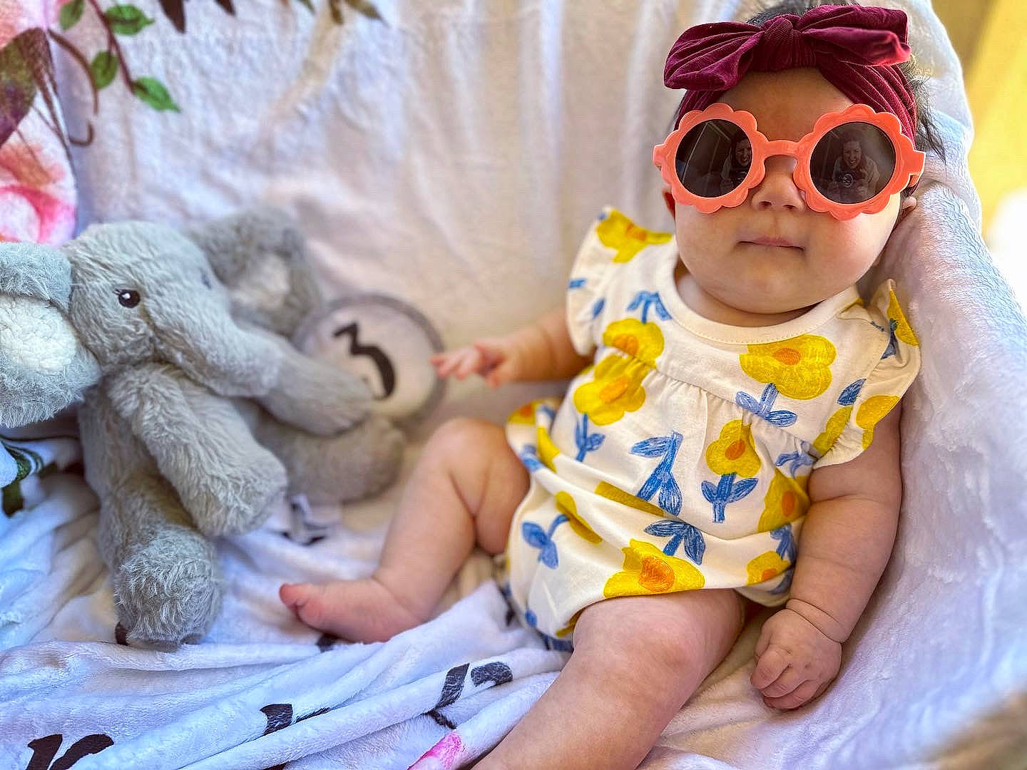 Georgia is registered to the contest to win money with this photo: baby, baby_toddler_clothing, cap, doll, dress, eyewear, fawn, finger, glasses, goggles, hand, happy, headgear, headwear, pattern, person, personal_protective_equipment, skin, sleeve, sunglasses