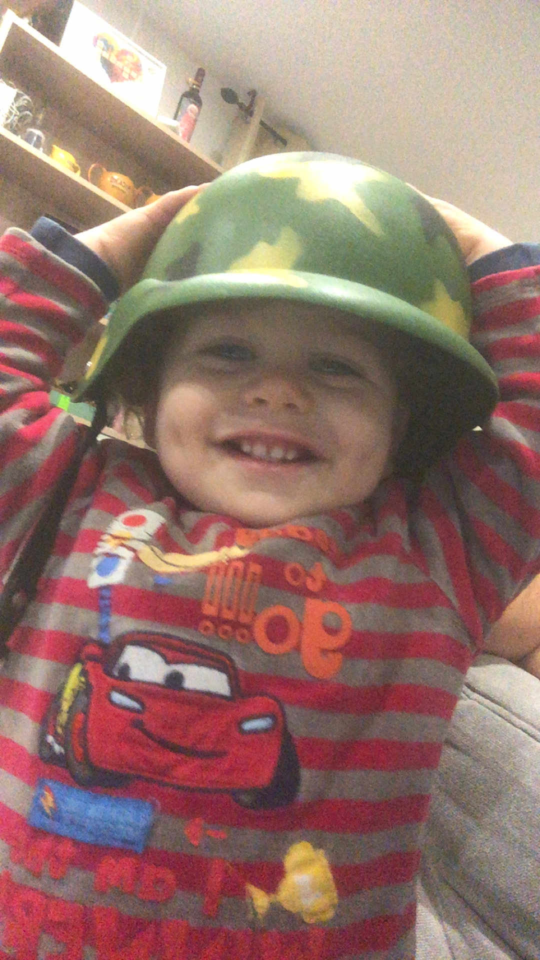 Mymy a rejoint le concours — aidez-le/la à gagner de superbes lots ! toddler, child, helmet, camouflage, striped_shirt, cartoon_car, smile, indoor, living_room, shelf, toy, happy, face, person, clothing, furniture, couch, cute, headwear, casual