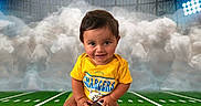 Blake is registered to the contest to win money with this photo: baby, child, football, sports, stadium, field, green_grass, yellow_shirt, smiling, cute, holding, sitting, sports_equipment, helmet_logo, clouds, lights, portrait, outdoor, young_child, person