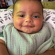 Blake is registered to the contest to win money with this photo: baby, smiling, bib, face, child, indoor, pillow, pattern, chubby_cheeks, clothing, infant, happy, person, cute, closeup, portrait, soft_texture, hand, seated, background