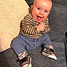 baby, smiling, sitting, couch, striped_clothing, converse_shoes, happy, indoor, furniture, cozy, hands_clasped, blue_pants, knitted_pillow, cute, child, portrait, head, footwear, person, expression