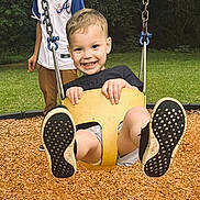 Kolton is registered to the contest to win money with this photo: child, swing, playground, smiling, feet, yellow_swing, grass, trees, person, outdoor, happy, young_child, wood_chips, casual_clothing, fun, park, daylight, play, active, joy