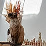 cat, siamese_cat, wooden_table, dried_grass, chess_set, cat_figurine, indoor, decor, still_life, pets, animal, feline, brown_cat, ornamental_plants, tabletop, sitting, side_profile, quiet, calm, natural_light