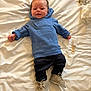 baby, blue_hoodie, curious_text, bed, white_bedspread, small_brush, tissue, beige_shoes, dark_pants, infant, lying_down, indoors, cute, child, person, smiling, cozy, casual_clothing, newborn, soft_lighting
