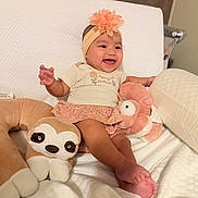 Samantha joined the competition — help win amazing prizes! baby, child, smiling, sitting, headband, flower, plush_toy, stuffed_animal, sloth, pink_toy, bed, bedding, pillow, indoor, portrait, infant, happy, clothing, floral_skirt, nursery
