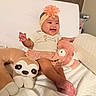 baby, child, smiling, sitting, headband, flower, plush_toy, stuffed_animal, sloth, pink_toy, bed, bedding, pillow, indoor, portrait, infant, happy, clothing, floral_skirt, nursery