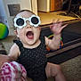 Ava is registered to the contest to win money with this photo: baby, child, sunglasses, glasses, flower_shaped, black_clothing, indoor, carpet, toy, hand, mouth_open, blurred_motion, cheerful, person, living_room, floor, colorful, furniture, door, playful