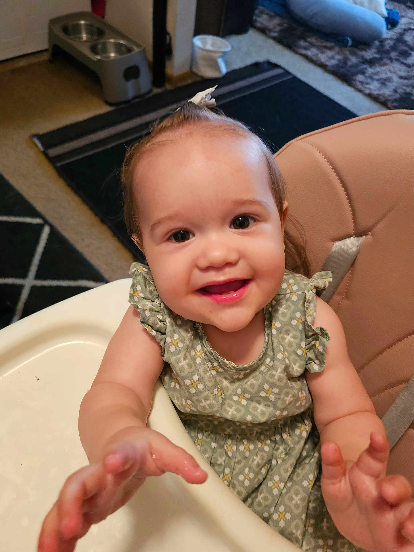 Ava is registered to the contest to win money with this photo: toddler, child, high_chair, smiling, happy, indoor, baby, clothing, dress, face, hands, hair, bow, kitchen, carpet, furniture, person, seat, table, cute