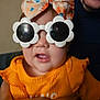 child, sunglasses, headband, orange_clothing, baby, person, indoor, adult_hand, face, accessory, portrait, cute, fashion, colorful, smile, holding, glasses, baby_clothes, closeup, fun