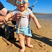 Nino participe au concours pour gagner de l'argent avec cette photo : baby, child, sunglasses, beach, sand, sunny, summer, person, toddler, blue_sky, stroller, hands, clothing, shorts, white_shirt, outdoor, foot, smile, playful, vacation