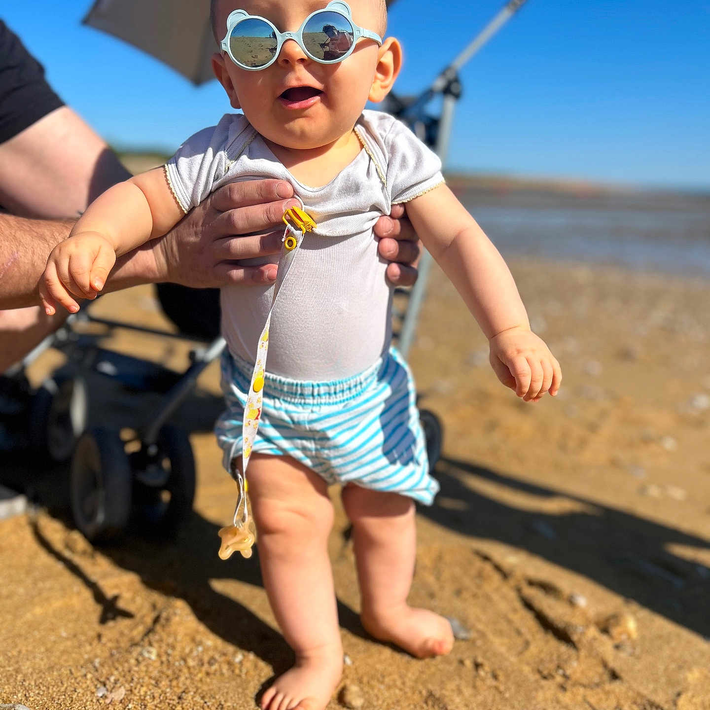 Nino participe au concours pour gagner de l'argent avec cette photo : baby, child, sunglasses, beach, sand, sunny, summer, person, toddler, blue_sky, stroller, hands, clothing, shorts, white_shirt, outdoor, foot, smile, playful, vacation