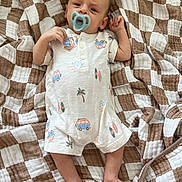 Crew is registered to the contest to win money with this photo: baby, infant, blanket, onesie, pacifier, face, foot, hand, lying_down, pattern, brown, white, clothing, person, cute, sleepwear, newborn, indoors, soft, comfort