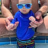 Crew is registered to the contest to win money with this photo: baby, child, sunglasses, blue_shirt, swim_shorts, pool, water, adult_hands, bare_chest, summer, outdoor, sunny, colorful, protective, playful, footwear_none, person, fashion, casual, fun