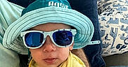 Crew joined the competition — help win amazing prizes! toddler, child, sunglasses, hat, yellow_shirt, couch, pillow, indoor, casual, cute, portrait, baby, relaxing, smiling, clothing, blue, person, furniture, accessory, comfort