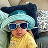 toddler, child, sunglasses, hat, yellow_shirt, couch, pillow, indoor, casual, cute, portrait, baby, relaxing, smiling, clothing, blue, person, furniture, accessory, comfort