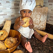 Isaiah is registered to the contest to win money with this photo: baby, child, chef_hat, bagel, bread, kitchen, countertop, flour, baking, cutting_board, rolling_pin, wooden_bowl, food, cooking, cute, apron, barefoot, person, indoors, snack