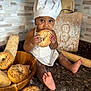 baby, child, chef_hat, bagel, bread, kitchen, countertop, flour, baking, cutting_board, rolling_pin, wooden_bowl, food, cooking, cute, apron, barefoot, person, indoors, snack