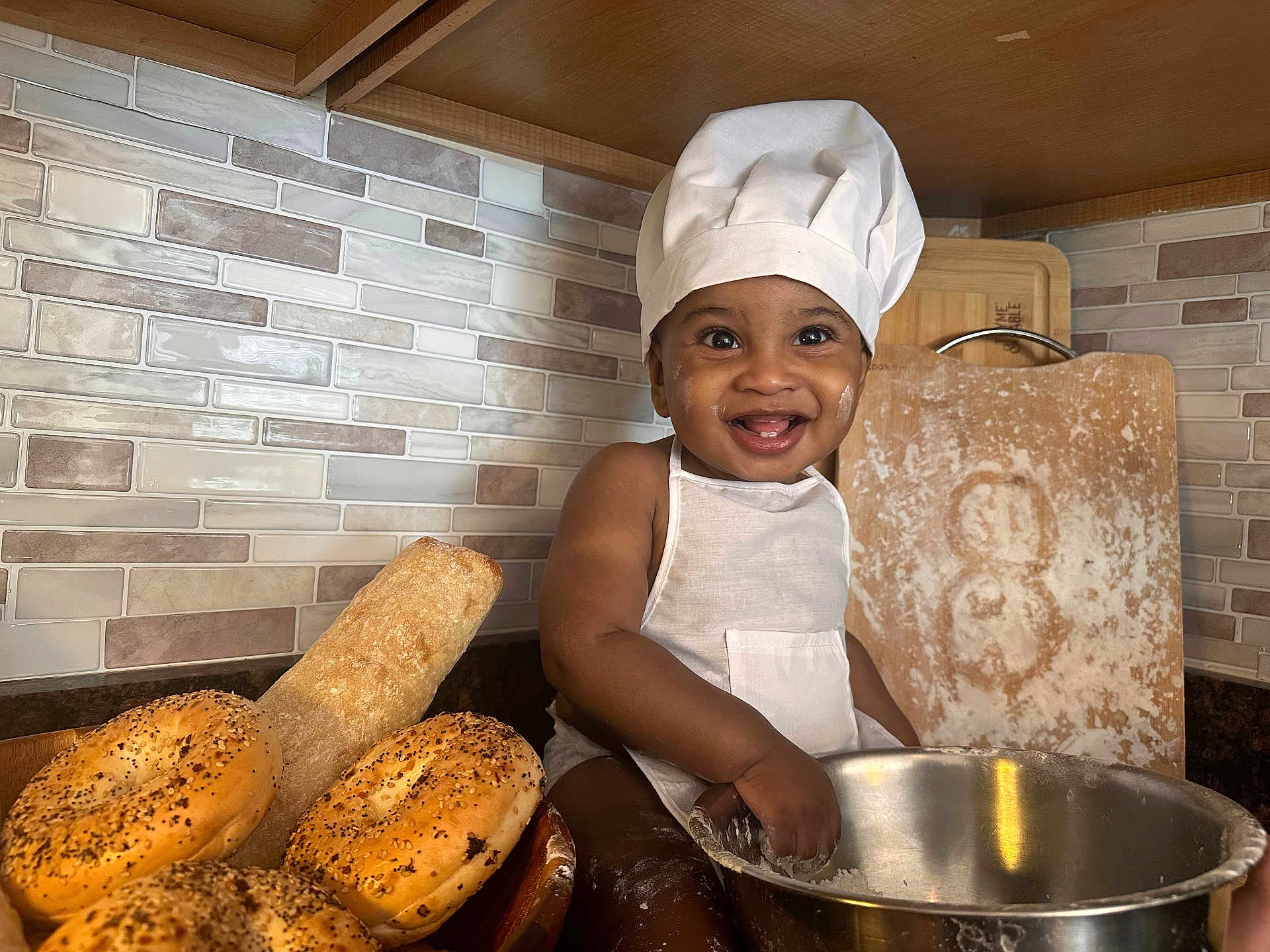 Isaiah joined the competition — help win amazing prizes! baby, child, chef_hat, apron, kitchen, baking, flour, bread, bagels, cutting_board, smile, happy, infant, metal_bowl, countertop, tile_wall, food, portrait, cooking, playful