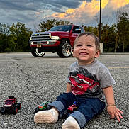 Oscar is registered to the contest to win money with this photo: toddler, child, toy_cars, red_truck, parking_lot, asphalt, cloudy_sky, trees, casual_clothing, socks, bracelet, happy, smiling, outdoor, vehicle, nature, playful, jeans, street_lamp, daylight