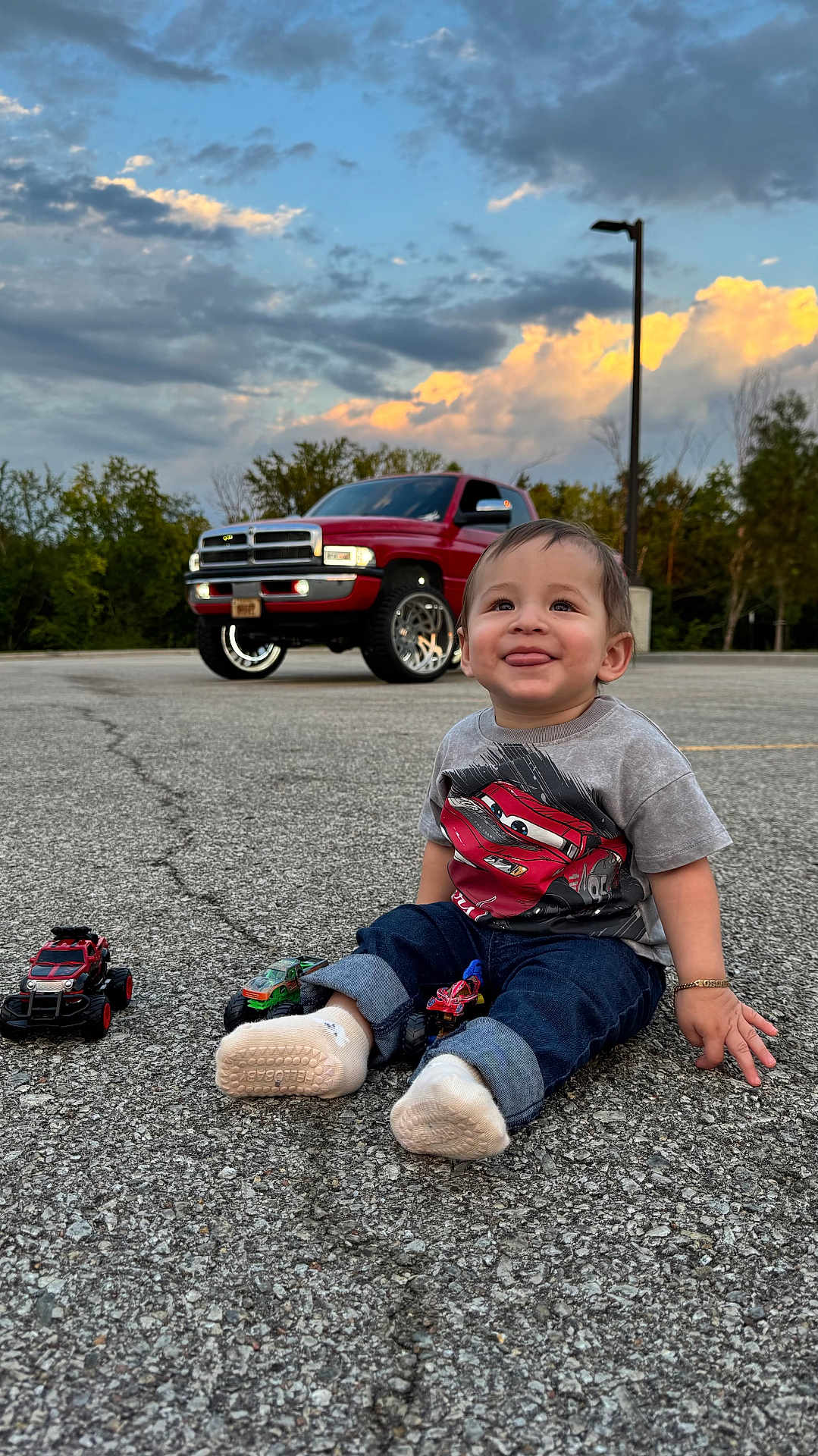 Oscar is registered to the contest to win money with this photo: toddler, child, toy_cars, red_truck, parking_lot, asphalt, cloudy_sky, trees, casual_clothing, socks, bracelet, happy, smiling, outdoor, vehicle, nature, playful, jeans, street_lamp, daylight