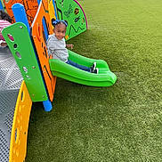 Isolyn is registered to the contest to win money with this photo: child, playground, slide, green_slide, artificial_turf, fence, blue_bows, gray_hoodie, outdoor, play_equipment, colorful, happy, smiling, person, recreation, fun, kids_play, daylight, casual_clothing, park