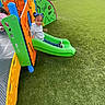 child, playground, slide, green_slide, artificial_turf, fence, blue_bows, gray_hoodie, outdoor, play_equipment, colorful, happy, smiling, person, recreation, fun, kids_play, daylight, casual_clothing, park