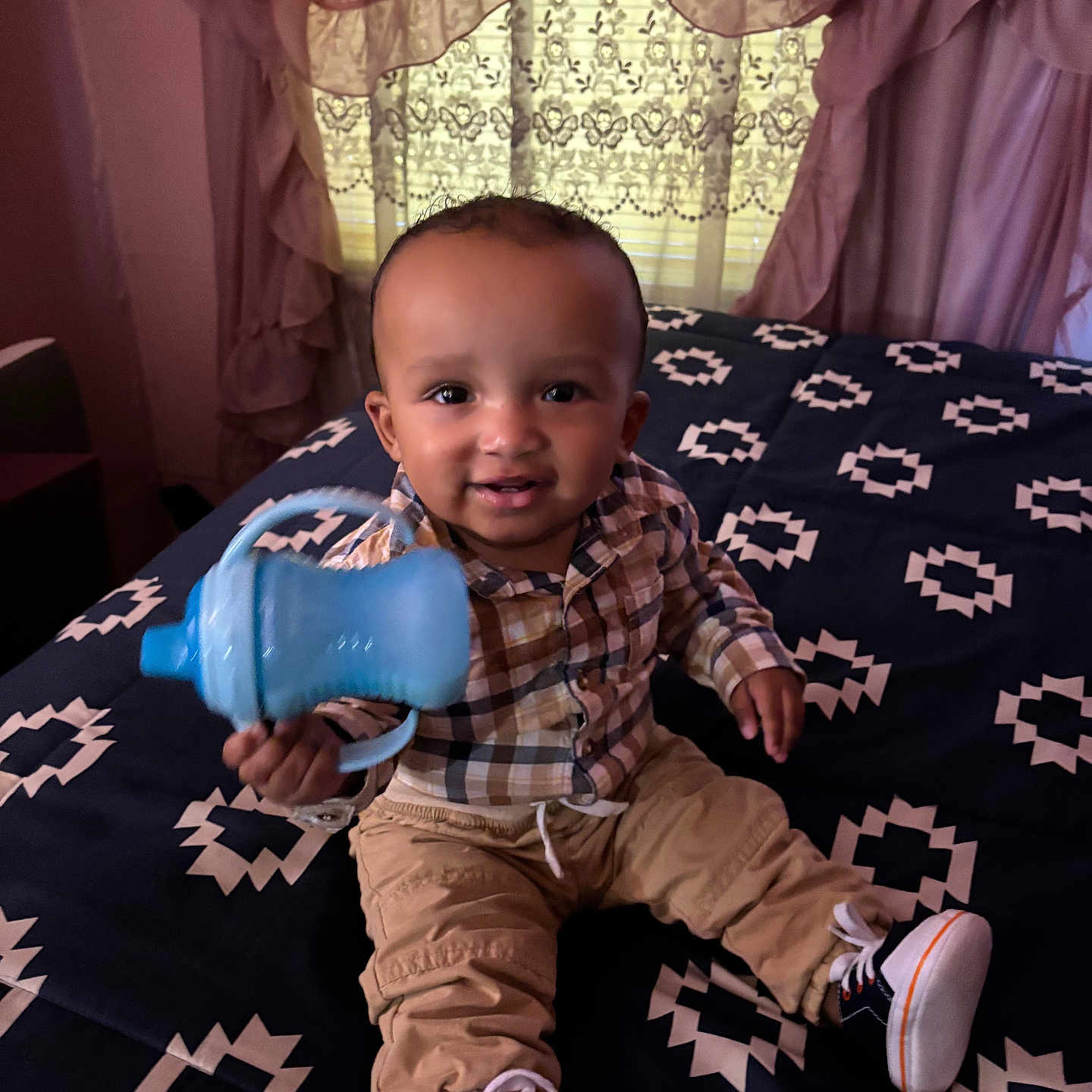 Akari-Reece is registered to the contest to win money with this photo: baby, bed, bedspread, child, curtains, face, furniture, happy, home, indoor, pants, pattern, person, plaid_shirt, seated, sippy_cup, smiling, sneakers, toddler, window