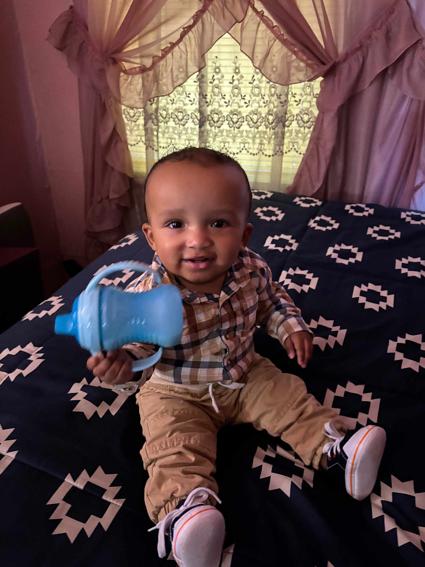 Akari-Reece is registered to the contest to win money with this photo: toddler, child, baby, sippy_cup, bed, bedspread, plaid_shirt, pants, sneakers, indoor, curtains, window, smiling, person, furniture, home, pattern, happy, face, seated