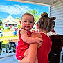 Remi is registered to the contest to win money with this photo: baby, child, toddler, red_clothing, hair_buns, porch, outdoor, daytime, family, holding, playful, tongue_out, smiling, blue_sky, house, grass, people, chair, summer, sunny
