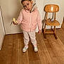 child, girl, banana, pink_jacket, hair_bun, hair_clips, indoor, wooden_floor, wooden_chair, sneakers, pants, playful_expression, standing, peeling_banana, cozy_clothing, white_wall, bag, flooring, furniture, home