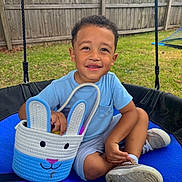 Malakai is registered to the contest to win money with this photo: child, boy, swing, outdoor, grass, wooden_fence, basket, bunny, toy, blue, smiling, shoes, shorts, tshirt, play, happy, face, person, spring, fun