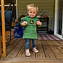 toddler, child, hoodie, green_clothing, jeans, socks, shoes, porch, wooden_floor, smile, blonde_hair, holding_objects, balls, home, chair, water_jugs, rug, doorway, outdoor, person
